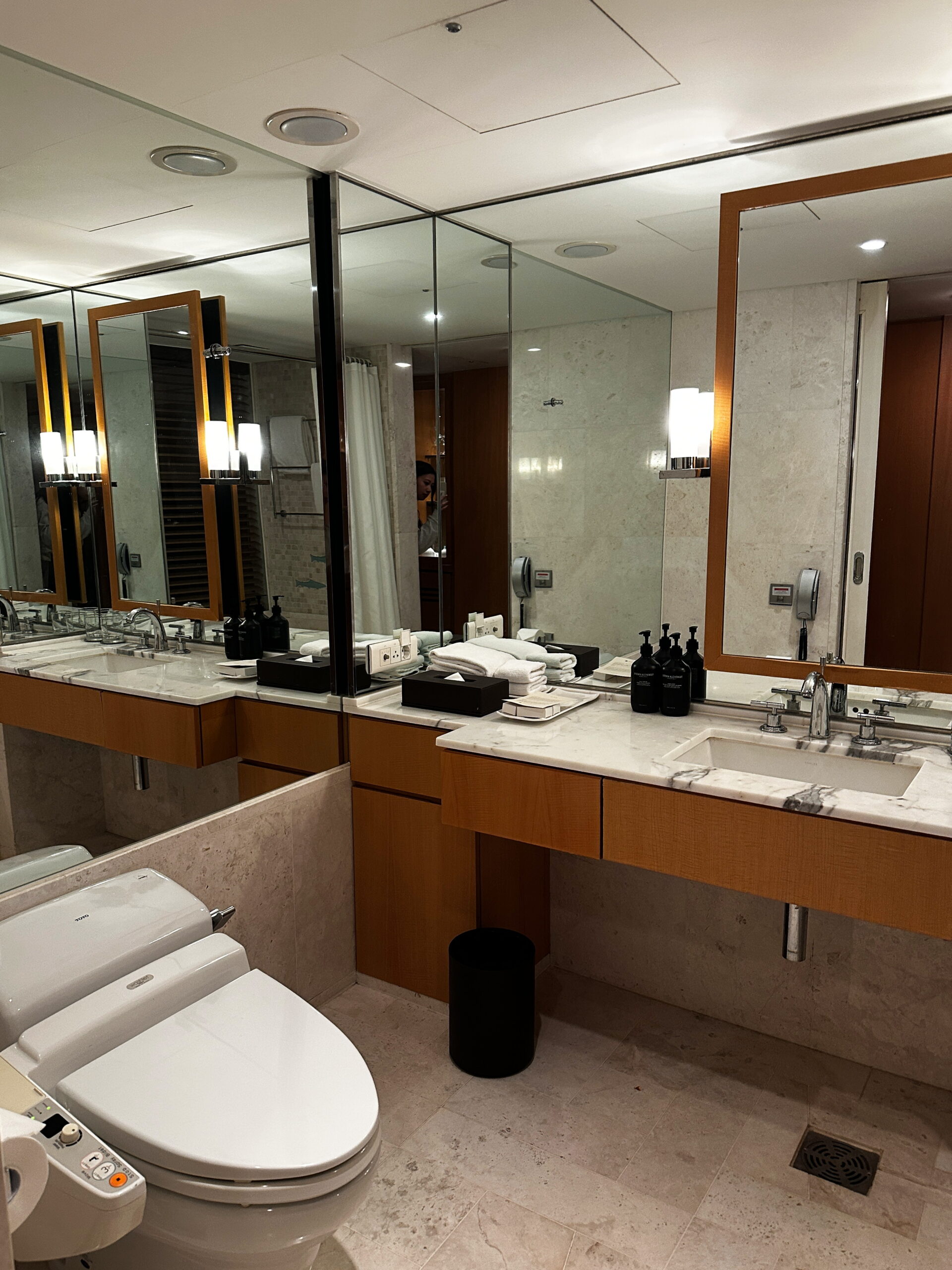 Elegant hotel bathroom featuring mirrors on both sides, creating a spacious feel. A sleek countertop with a single sink holds neatly arranged amenities. The toilet includes a bidet for added comfort and convenience.