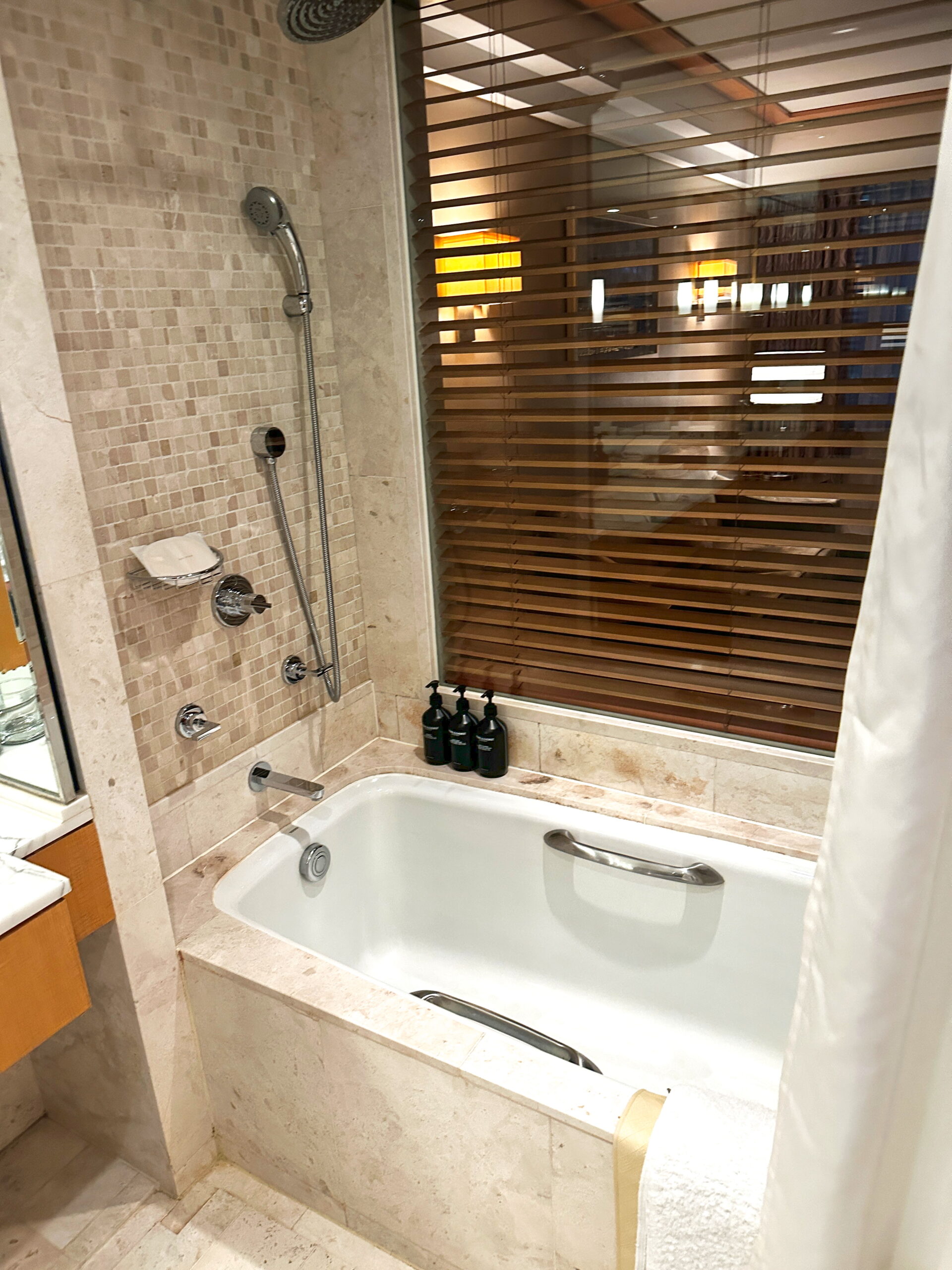 Bathroom in a Korean hotel featuring a bathtub with safety handles for accessibility, along with a detachable showerhead for convenience. Shampoo, conditioner, and body wash are provided. The tub is positioned next to a window with adjustable blinds for privacy.