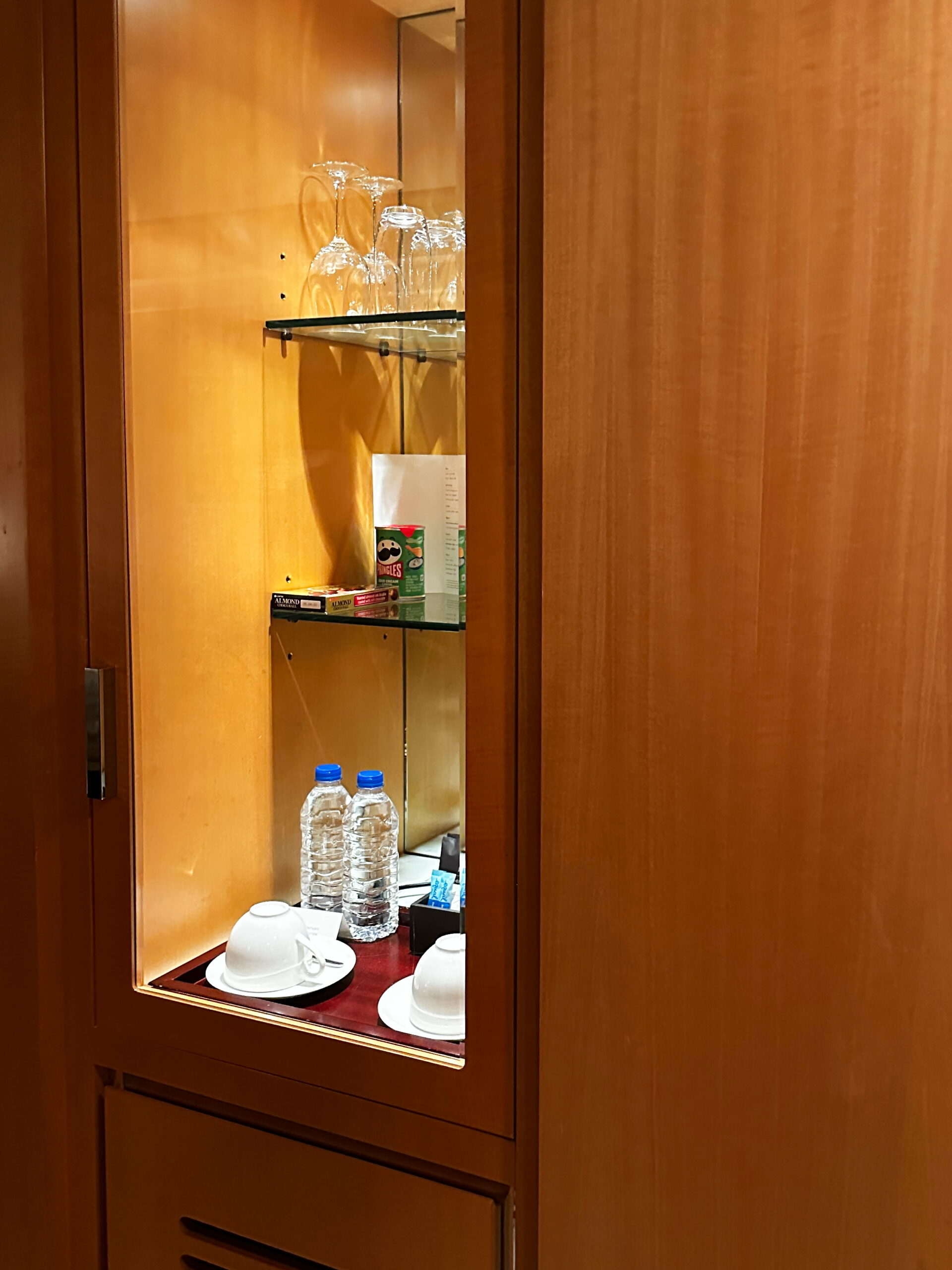Korean Hotel room cabinet with a mini fridge tucked underneath, storing complimentary water bottles and extra tea/coffee glasses with white saucers. Wine glasses are neatly arranged above, while additional amenities like toothpaste and toothbrushes are available for purchase, offering convenience during the stay.
