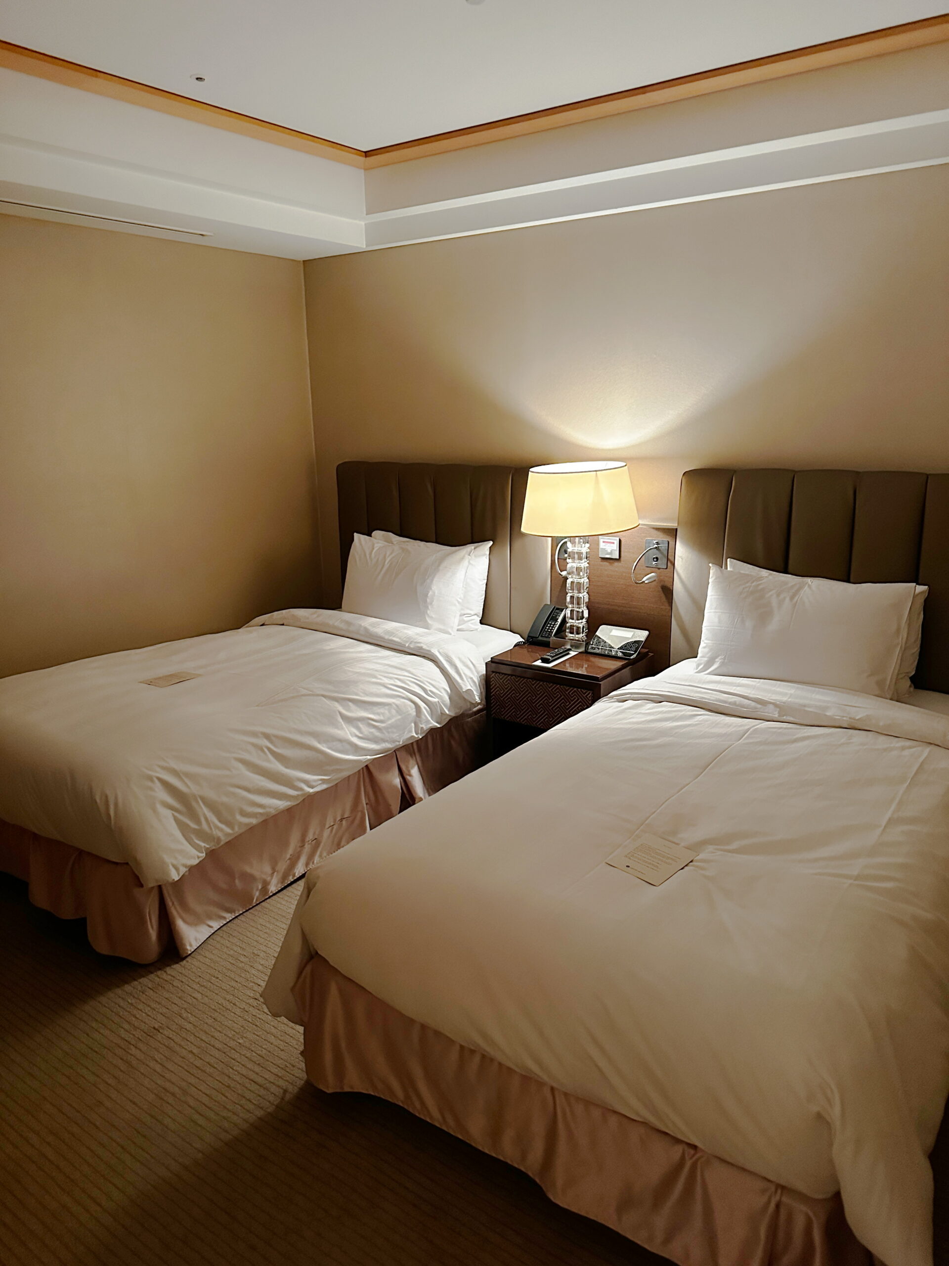 Korean Hotel room with two oversized twin beds, neatly made with plush white linens. A wooden nightstand sits in the middle, holding a softly glowing nightlamp that casts warm, cozy light upwards. A flat-screen TV is mounted on the wall, adding to the room's refined and comfortable feel.