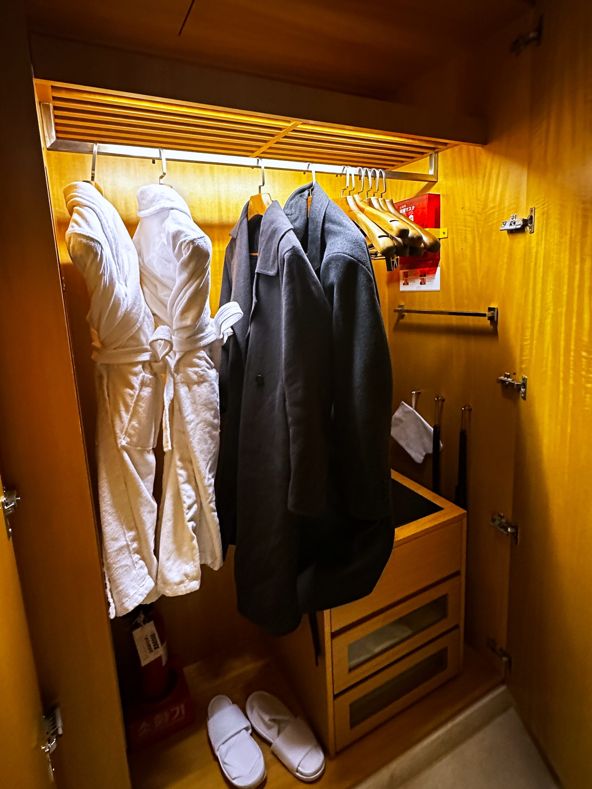 Elegant closet with oak shelving, offering white robes for guest use during the stay. A lockbox is provided for security, while the lower section provides space for shoes or a small suitcase. Small pull-out drawers offer extra storage, and built-in lighting illuminates the closet when in use.