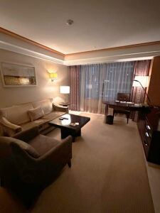 Elegant and cozy Korean hotel suite living room featuring a plush sectional couch, a single armchair, and a refined coffee table at the center. A beautifully framed picture adorns the wall, enhancing the warm ambiance. A classic wooden desk with a comfortable chair and soft task lighting creates an inviting workspace. A flat-screen TV is positioned nearby, while sliding drape curtains offer a glimpse of the hotel's front entrance, adding to the roomβs refined charm.